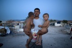 Roma young father came from Albania to Athens center shantytown to survive with his family. He holds his two babies at his arms. Shantytown in the background
 © Maro Kouri
